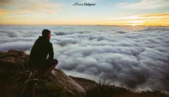 Man sitting on the top of a mountain overlooking a skyline with clouds as the sunsets